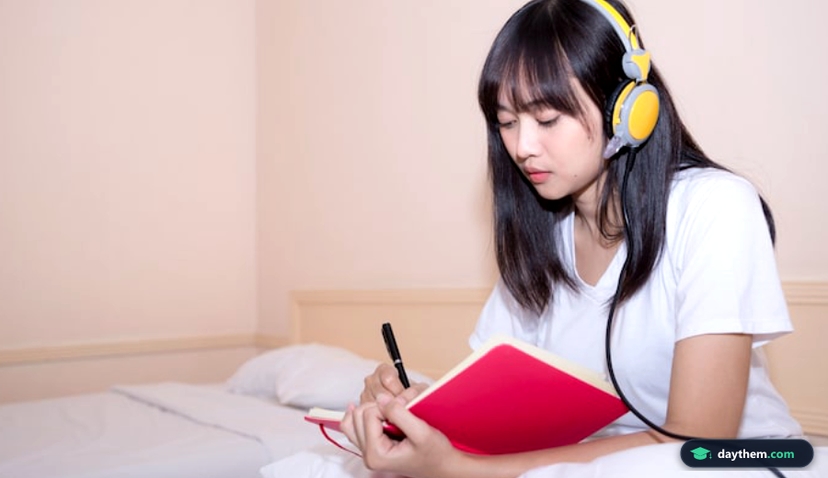 Một bạn trẻ mặc đồ ngủ ngồi trên giường đang viết vào sổ tay khi tự học tiếng Nhật tại nhà